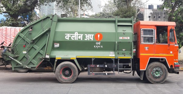 A green refuse compactor truck with an orange cab, marked 'Clean Up', parked on a Mumbai street
